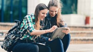 two young girls looking at ipad and laughing while sitting on the stairs outdoor | Trendy And Cute Outfits For School You Can Wear Every Day | Featured