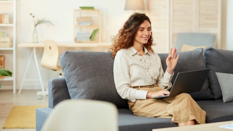 Young adult woman sitting in sofa at home participating in video conference, greeting her colleagues | Grooming Tips On How To Dress For Your Next Video Conference | what to wear to conference | Featured