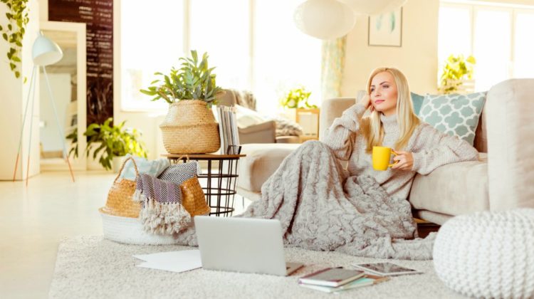 Beautiful young woman wearing sweater sitting on carpet in living room covered with blanket and resting with cup of coffee | Cute Throw and Fleece Blankets For A Warm and Productive Work From Home Experience | blanket shawl | Featured